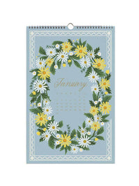 A spiral-bound January wall calendar featuring a floral wreath design in yellow, white, and green on a dusty blue background, with the word 'January' in gold script.