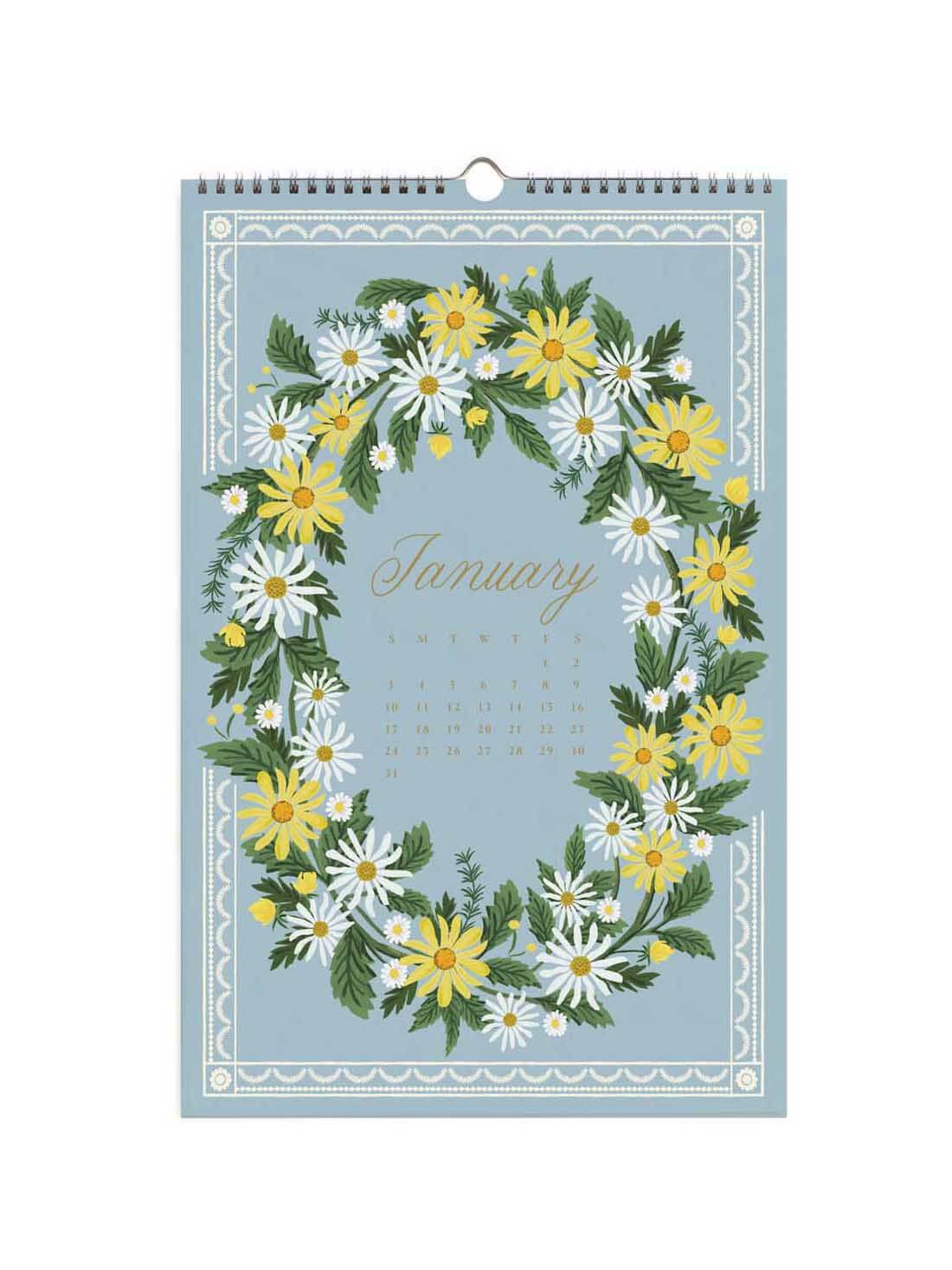 A spiral-bound January wall calendar featuring a floral wreath design in yellow, white, and green on a dusty blue background, with the word 'January' in gold script.