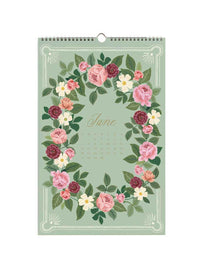 A hanging wall calendar page for June with a floral wreath design in pink, burgundy, cream, and green on a pale sage background. The word 'June' is printed in the center, with a small calendar grid below it.