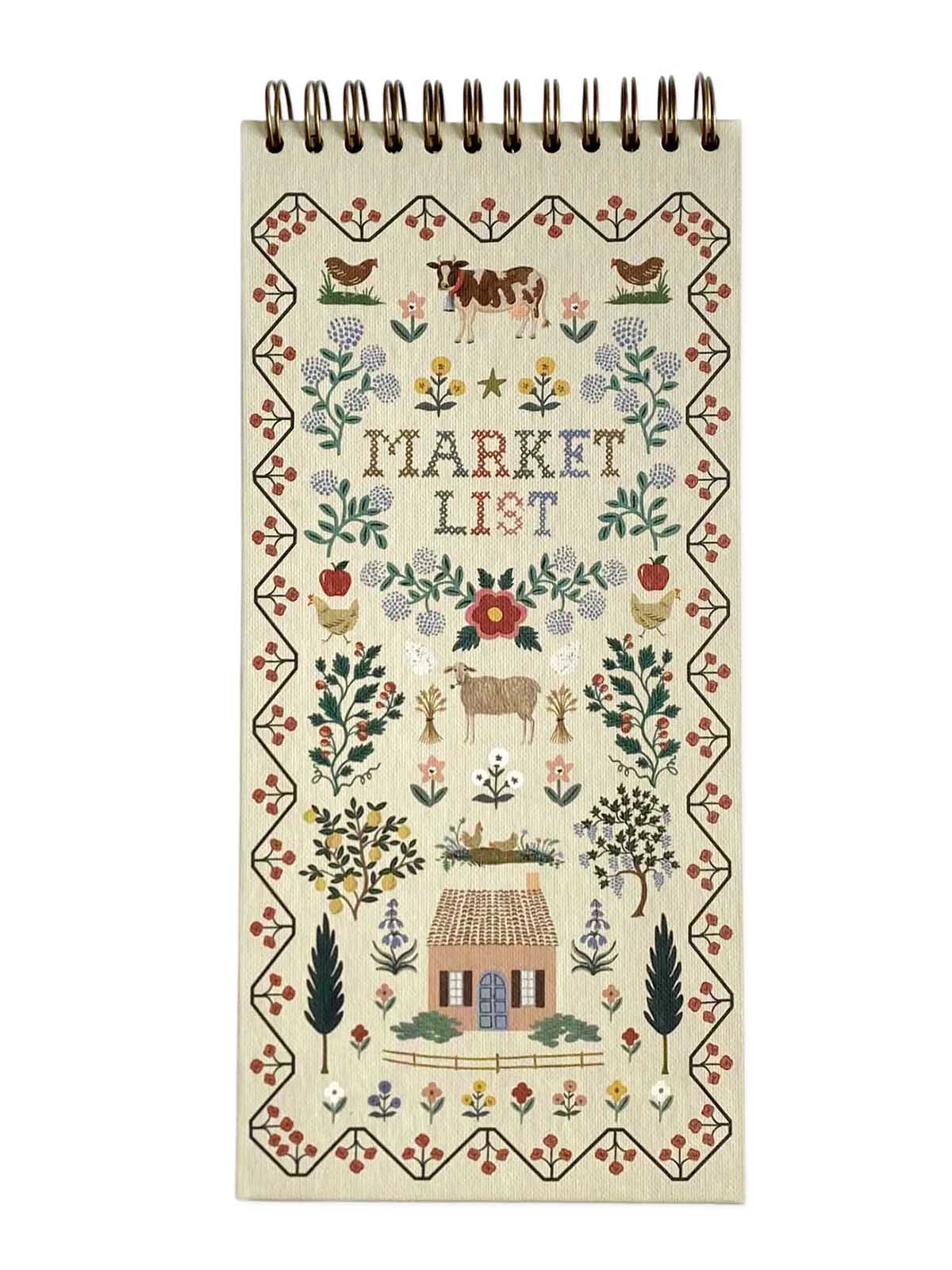 A spiral-bound notebook with a cream-colored cover featuring a detailed folk-art style cross-stitch design. The design includes the words 'MARKET LIST' in the center, surrounded by flowers, birds, a cow, a goat, a small house, and trees.
