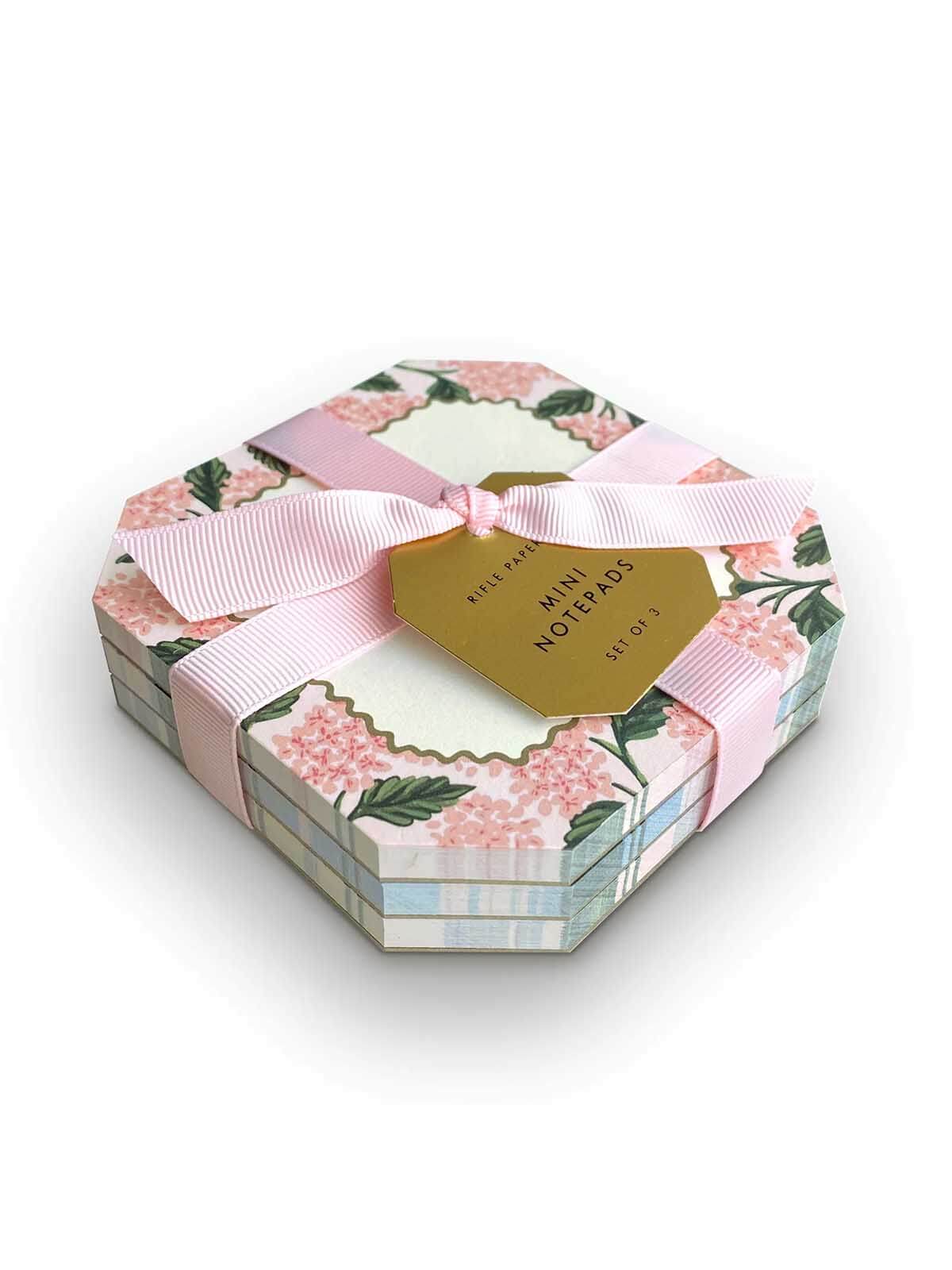 A stack of mini notebooks in a hexagonal gift box with a pink floral pattern, tied with a pink ribbon and a gold tag that reads 'Mini Notebooks'.